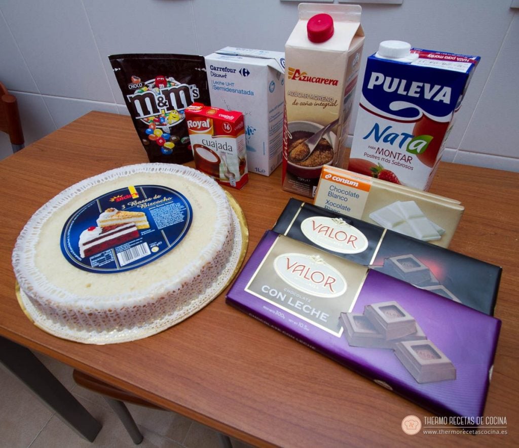 Los ingredientes sobre la mesa: la plancha de bizcocho comprada, los sobres de cuajada, la leche, el azúcar, el brik de nata para montar, las tabletas de chocolate negro, con leche y blanco, y una bolsa de M&amp;M's