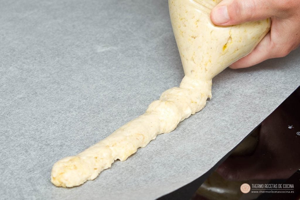 masa de croqueta saliendo de una bolsa cortada por la punta, formando un cordón sobre papel de horno