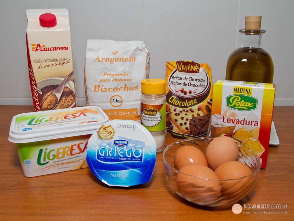 Bizcocho con pepitas de chocolate | 3 | Recetas de cocina para Thermomix Ingredientes del bizcocho alineados sobre la mesa: brik de azúcar moreno de caña, harina para bizcochos, caja de perlitas de chocolate, aceitera, caja de levadura, tarrina de margarina, un yogur griego natural, azúcar avainillado y cuatro huevos en un cuenco
