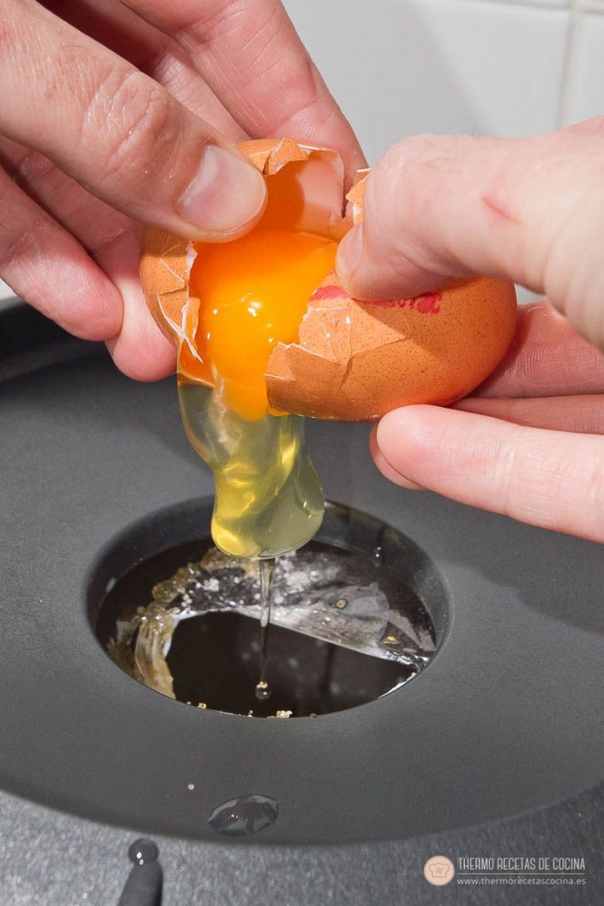 Natillas de napolitana | 8 | Recetas de cocina para Thermomix dos manos cascando un huevo por el bocal de la tapa de la Thermomix, con la yema cayendo dentro del vaso