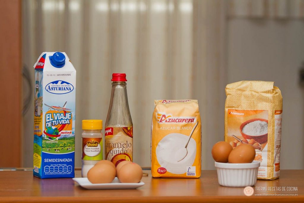 Ingredientes del bizcoflan alineados sobre la mesa: brik de leche semidesnatada, botecito de azúcar avainillado, botella de caramelo líquido, paquete de azúcar blanco, harina y los huevos repartidos en un plato y un cuenco