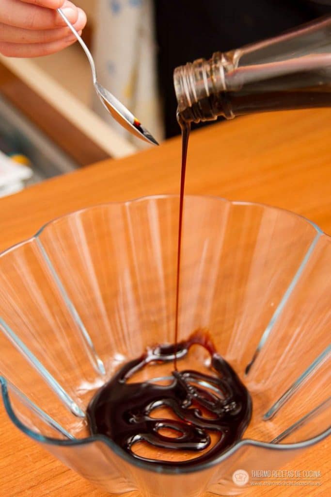 Botella de caramelo líquido vertiendo un hilo de caramelo en el fondo de un molde de cristal acanalado, con una cuchara al lado