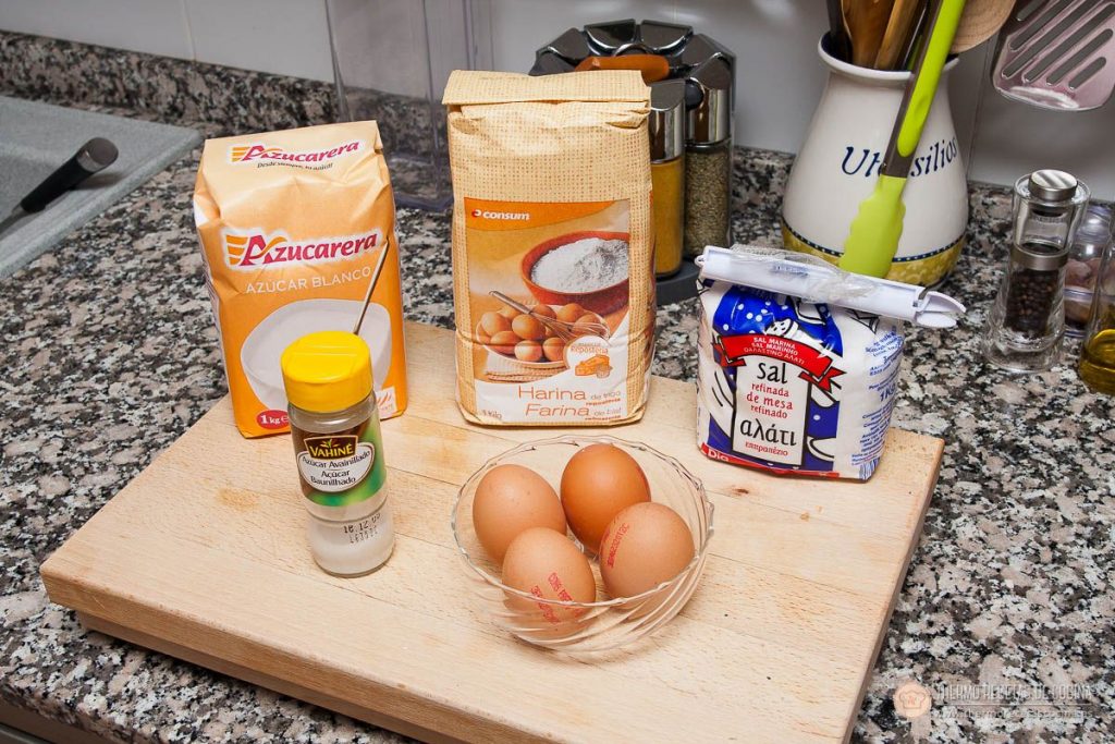 azúcar, harina de repostería, un botecito de azúcar avainillado, sal y tres huevos en un cuenco de cristal sobre una tabla de madera, los ingredientes del bizcocho genovés