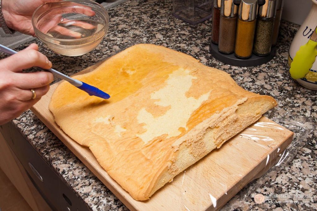 humedeciendo el bizcocho genovés extendido con almíbar, con un pincel de silicona y un cuenco de almíbar al lado
