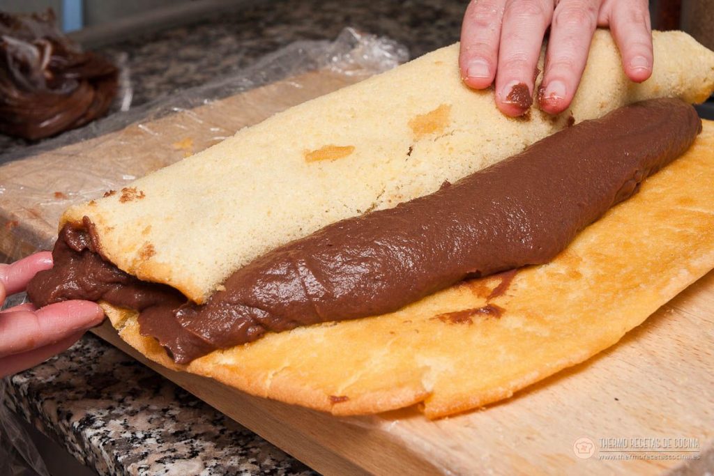 las dos manos levantando el bizcocho para enrollarlo sobre la tira de trufa de chocolate