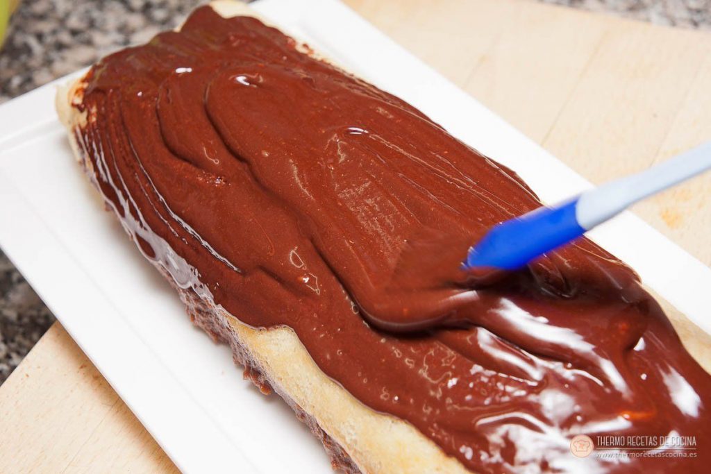 extendiendo con una espátula la cobertura de chocolate brillante sobre el tronco, ya colocado en la fuente blanca