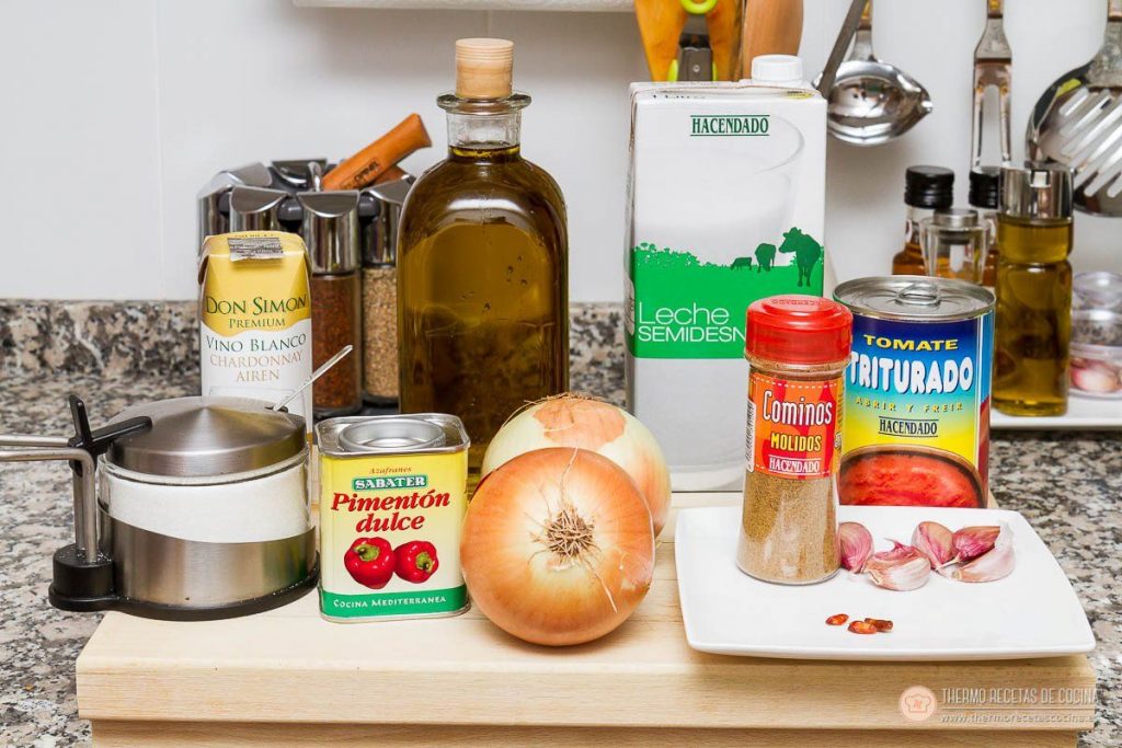 Salsa brava en thermomix | 3 | Thermo Recetas Cocina los ingredientes de la salsa brava sobre la encimera: dos cebollas, dientes de ajo y guindilla en un plato, lata de tomate triturado, brik de leche, botella de vino blanco, aceite, azucarero, comino molido y una lata de pimentón dulce
