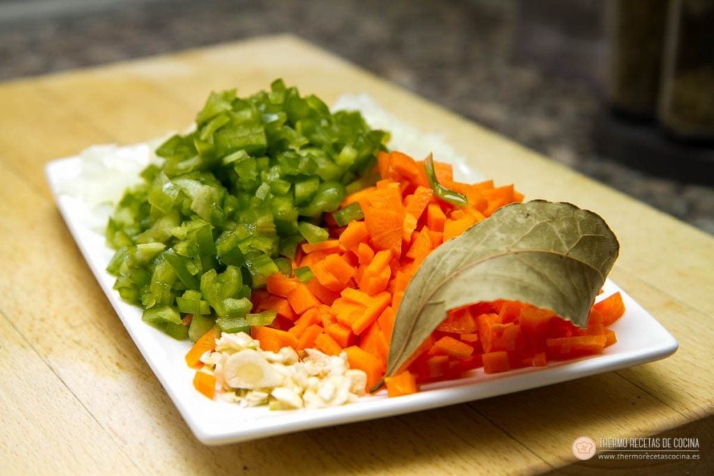 Pimiento verde, zanahoria, cebolla y ajo picados sobre una fuente blanca, con la hoja de laurel encima