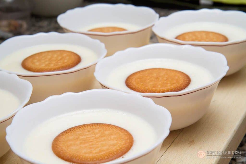 Natillas | 10 | Recetas de cocina para Thermomix Seis cuencos blancos de natillas con su galleta María en el centro, alineados y listos para la nevera