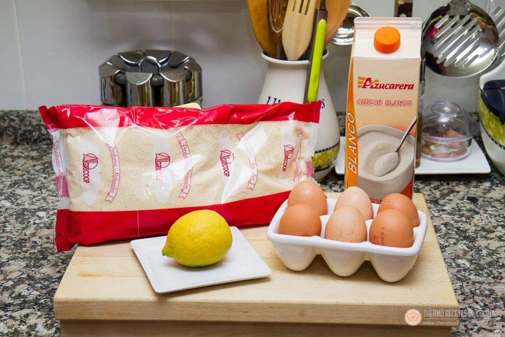 ingredientes de los almendrados sobre una tabla de madera: bolsa de almendra molida, brik de azúcar, un limón y una huevera con seis huevos