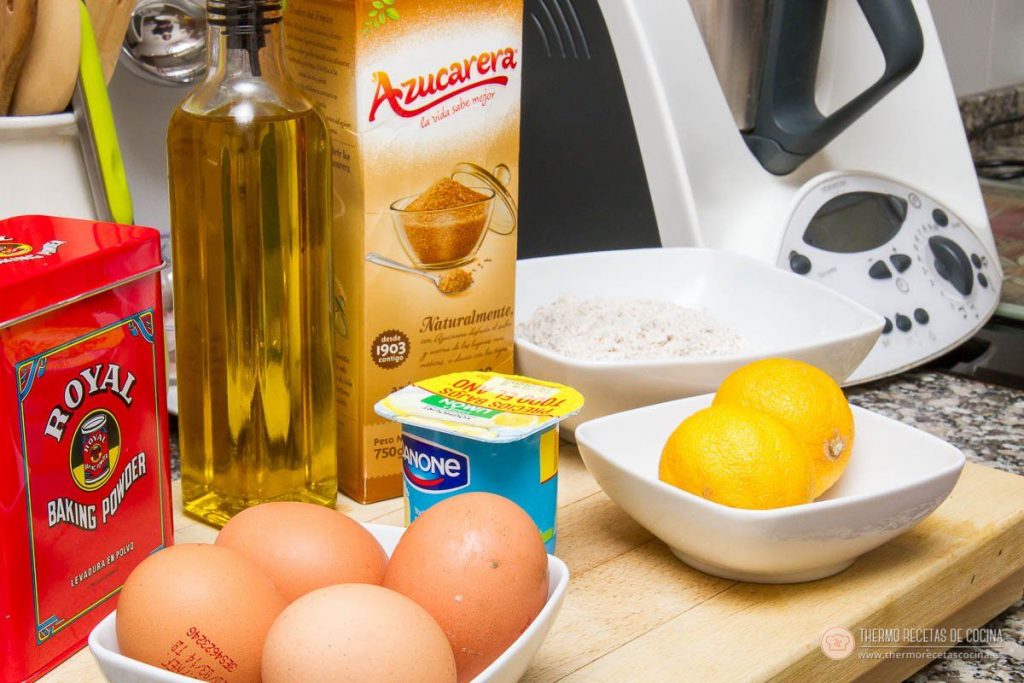ingredientes del bizcocho de espelta sobre la encimera con la Thermomix detrás: aceite, levadura Royal, azúcar moreno, harina de espelta, yogur, cuatro huevos y dos limones