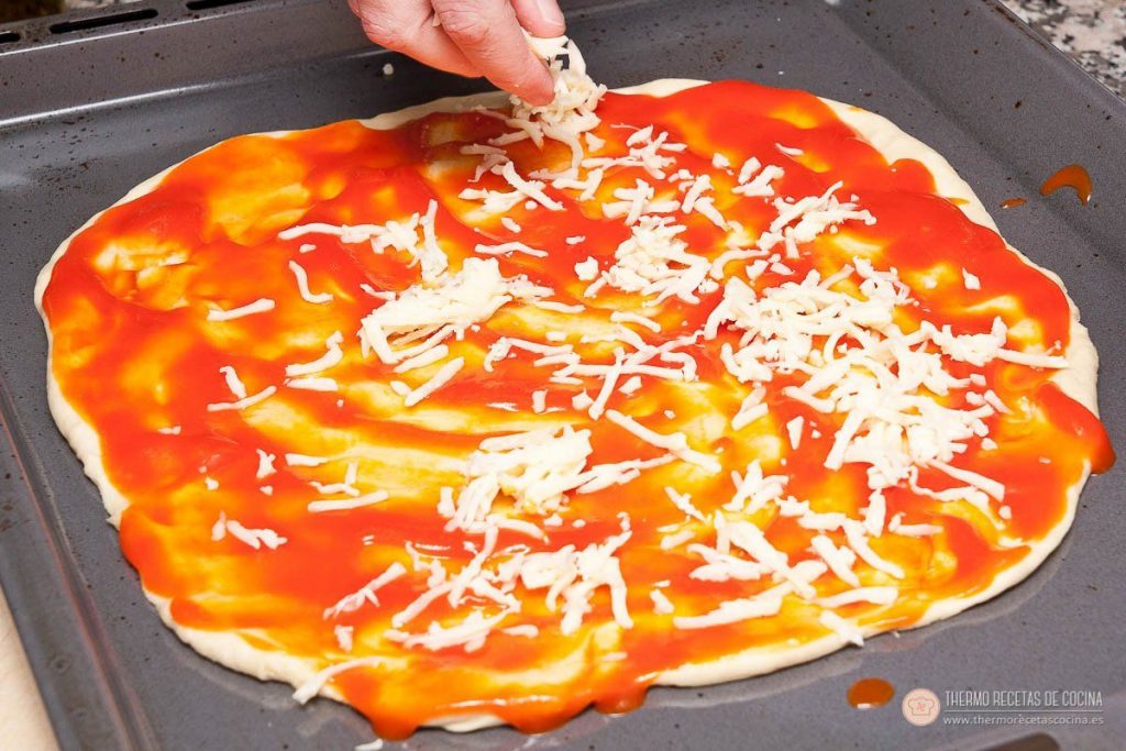 Base de pizza extendida en la bandeja, con el tomate ya repartido y la mano echando el queso rallado por encima
