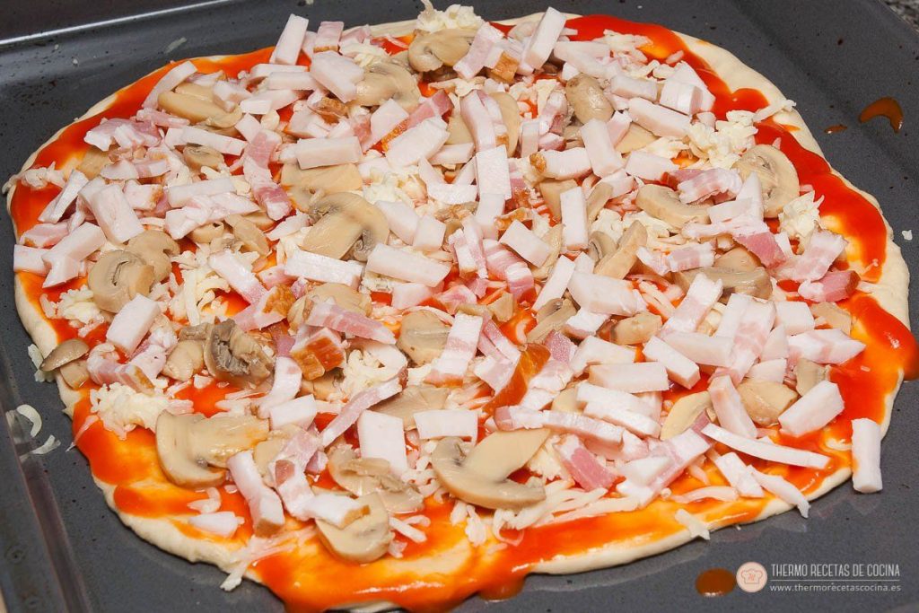 La pizza montada en la bandeja, con tomate, mozzarella rallada, champiñones laminados y tiras de beicon, lista para el horno
