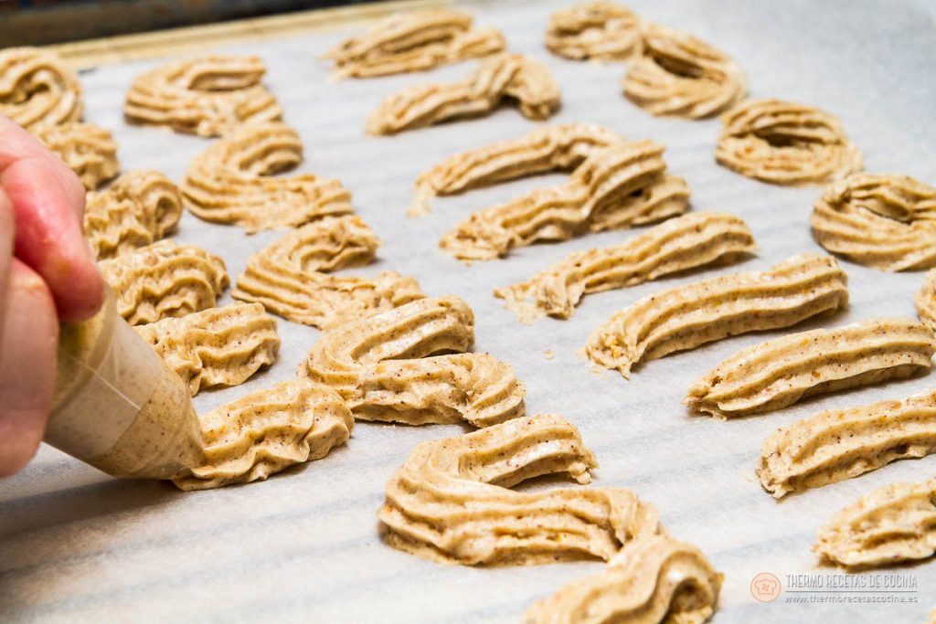 masa de las pastas saliendo de una manga pastelera con boquilla rizada, formando eses y tiras sobre el papel vegetal de la bandeja