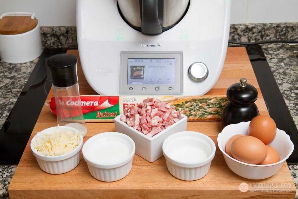 Receta de tarta Quiché de Bacon y Queso | 3 | Recetas de cocina para Thermomix los ingredientes de la quiche delante de la Thermomix: paquete de masa brisa, cuenco de dados de bacon, queso rallado, leche, nata, huevos y un molinillo de pimienta